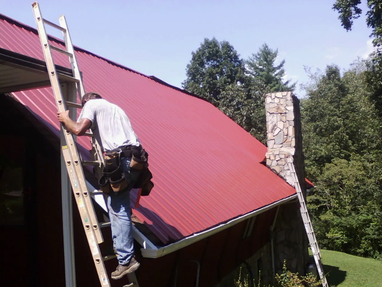 Roofer on a ladder beside a red metal roof and stone chimney — Soffit & Fascia Repair by Roofing Brothers in Hainesport